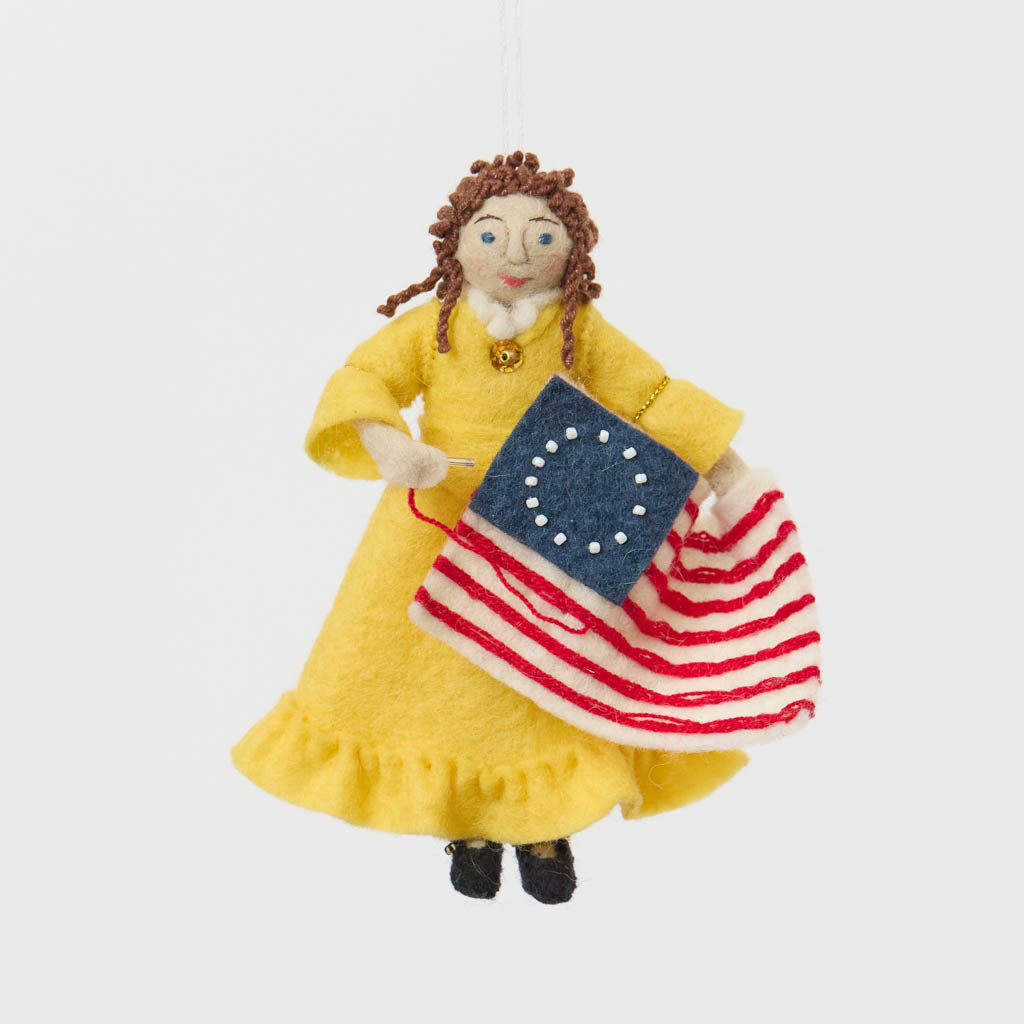 Celebrate the legendary maker associated with America’s first flag with our handcrafted Betsy Ross Ornament. Dressed in a bright colonial gown and holding the iconic early American flag, Betsy honors the spirit of independence, ingenuity, and handmade tradition.