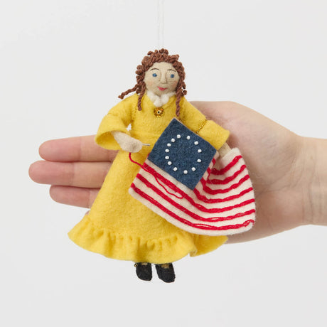 Celebrate the legendary maker associated with America’s first flag with our handcrafted Betsy Ross Ornament. Dressed in a bright colonial gown and holding the iconic early American flag, Betsy honors the spirit of independence, ingenuity, and handmade tradition.