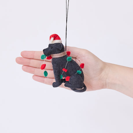 Craftspring’s Black Lab in Christmas Lights Ornament displayed in front of hand to show its size — felt dog wrapped in Christmas lights and wearing a Santa hat, a charming heirloom-quality piece for Christmas gifting — ethically crafted by artisans using sustainable wool.