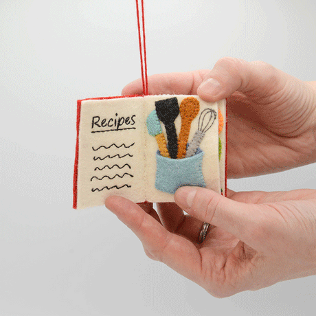 Handmade felt Bon Appetit Cookbook ornament from Craftspring shown flipping through interior pages—featuring intricate beaded and embroidered details inspired by cooking and recipes, ethically crafted by artisans using sustainable wool