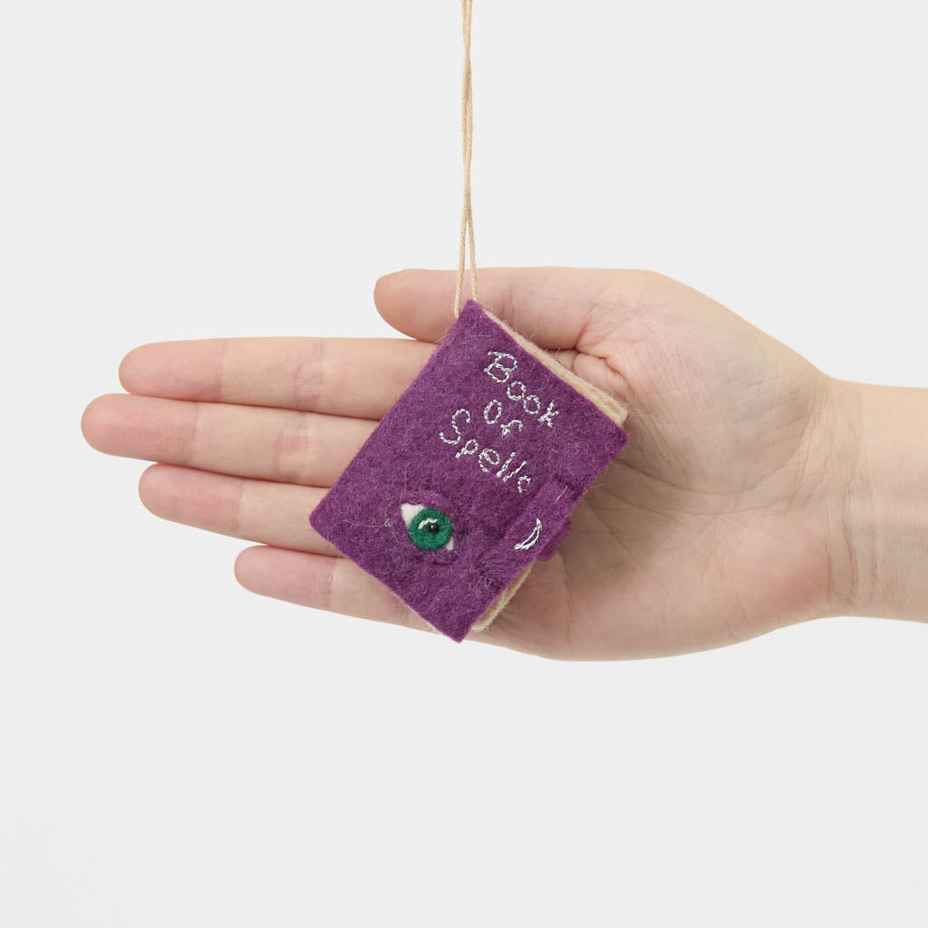 A handmade felt Book of Spells Ornament designed like a miniature mystical grimoire, complete with embroidered details, moon phases, herbal remedies, and a magical all-seeing eye.