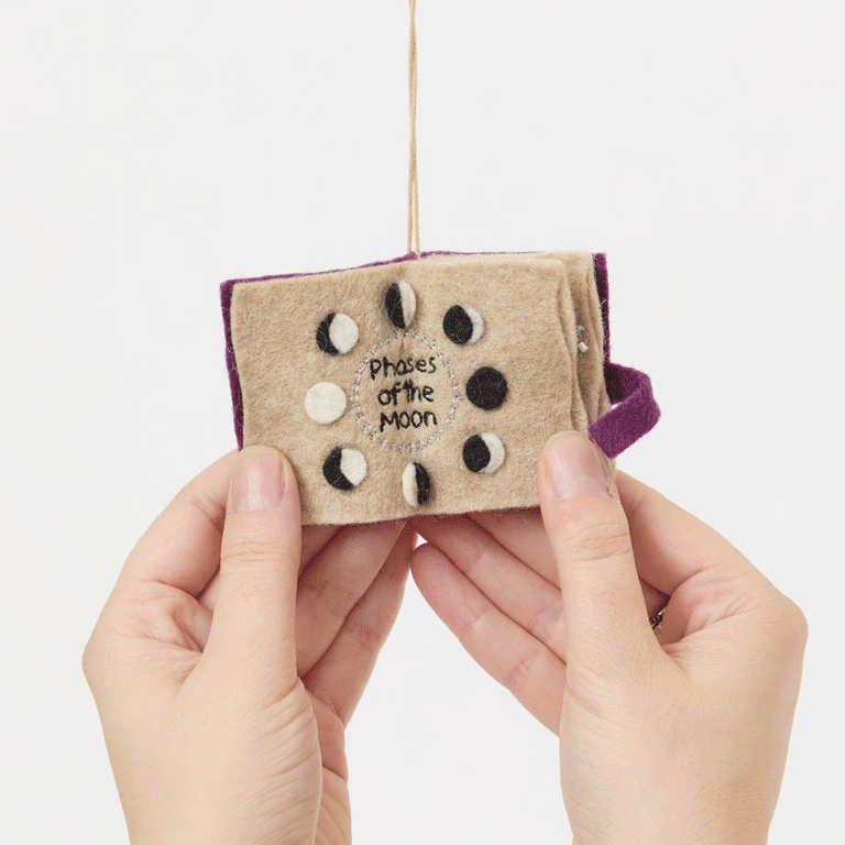 A handmade felt Book of Spells Ornament designed like a miniature mystical grimoire, complete with embroidered details, moon phases, herbal remedies, and a magical all-seeing eye.