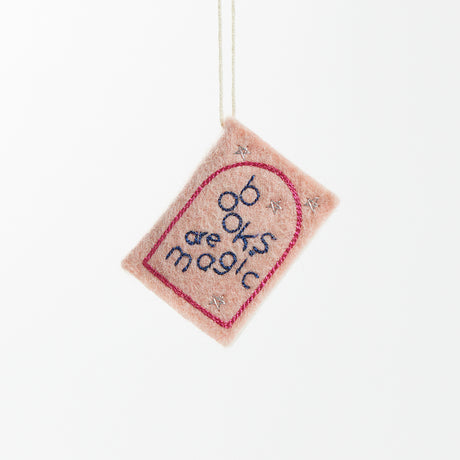 Hot pink felt ornament shaped like a small book, featuring delicately embroidered open pages. Inspired by the beloved Brooklyn bookstore Books Are Magic, this hand-felted wool ornament includes a hanging string and measures approximately 2.5 inches tall. A colorful tribute to the joy of reading and storytelling