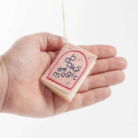 Hot pink felt ornament shaped like a small book, featuring delicately embroidered open pages. Inspired by the beloved Brooklyn bookstore Books Are Magic, this hand-felted wool ornament includes a hanging string and measures approximately 2.5 inches tall. A colorful tribute to the joy of reading and storytelling.  Held in a hand for scale reference.