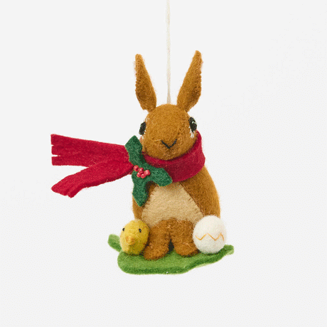 Our Brown Holiday Bunny with Chick Ornament is a sweet story of kindness and holiday cheer. Wrapped in a cozy crimson scarf adorned with felt holly and tiny red beads, this gentle brown bunny sits besides fluffy yellow chicks and a freshly hatched egg, capturing the magic of winter and the simple joy of being together.