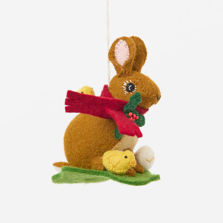 Our Brown Holiday Bunny with Chick Ornament is a sweet story of kindness and holiday cheer. Wrapped in a cozy crimson scarf adorned with felt holly and tiny red beads, this gentle brown bunny sits besides fluffy yellow chicks and a freshly hatched egg, capturing the magic of winter and the simple joy of being together.