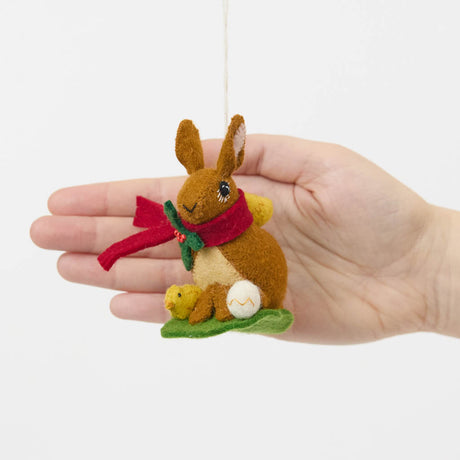 Our Brown Holiday Bunny with Chick Ornament is a sweet story of kindness and holiday cheer. Wrapped in a cozy crimson scarf adorned with felt holly and tiny red beads, this gentle brown bunny sits besides fluffy yellow chicks and a freshly hatched egg, capturing the magic of winter and the simple joy of being together.