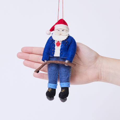 Craftspring’s Canadian Santa Ornament displayed in front of a hand to show its size — felt Santa dressed in a denim jacket and jeans with ice skates, holding a hockey stick, and featuring a Canadian flag maple leaf, a charming heirloom-quality piece for Christmas gifting — ethically crafted by artisans using sustainable wool.
