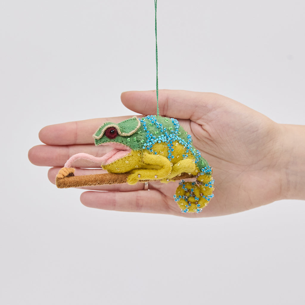 Craftspring’s Chameleon ornament held in front of a hand to show its size—handmade from sustainable wool with beaded details, a curled tail, and a long tongue catching a bug, ethically crafted by artisans using sustainable wool.