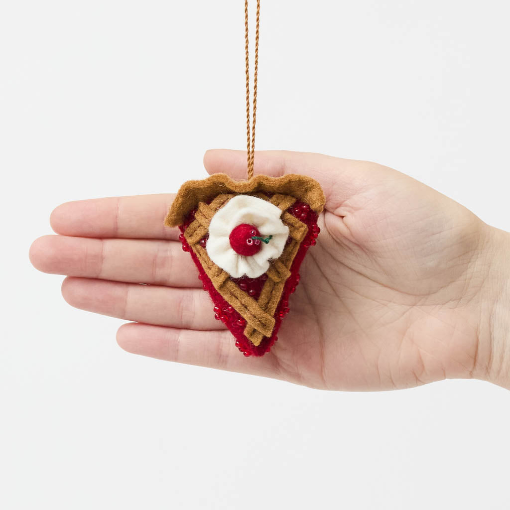 A true slice of nostalgia, our Cherry Pie Slice Ornament captures the timeless charm of one of America's favorite desserts. Handcrafted from natural wool felt, this delightful ornament features a rich cherry filling accented with sparkling red beadwork, a beautifully detailed lattice crust, a dollop of whipped cream, and a bright cherry on top.