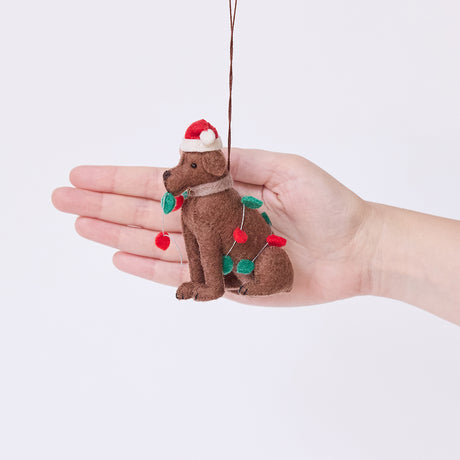 Craftspring’s Chocolate Lab in Christmas Lights Ornament displayed in front of hand to show its size — felt dog wrapped in Christmas lights and wearing a Santa hat, a charming heirloom-quality piece for Christmas gifting — ethically crafted by artisans using sustainable wool.