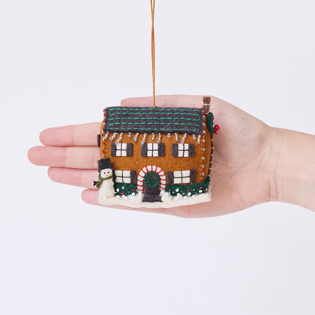 Craftspring’s Christmas House Ornament displayed in front of a hand to show its size — felt house with intricate beading, embroidery, and applique, including a snowman in the yard, beaded icicles, and holly bushes, a charming heirloom-quality piece for Christmas gifting — ethically crafted by artisans using sustainable wool.