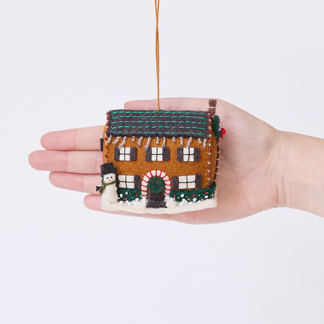 Craftspring’s Christmas House Ornament displayed in front of a hand to show its size — felt house with intricate beading, embroidery, and applique, including a snowman in the yard, beaded icicles, and holly bushes, a charming heirloom-quality piece for Christmas gifting — ethically crafted by artisans using sustainable wool.