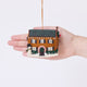 Craftspring’s Christmas House Ornament displayed in front of a hand to show its size — felt house with intricate beading, embroidery, and applique, including a snowman in the yard, beaded icicles, and holly bushes, a charming heirloom-quality piece for Christmas gifting — ethically crafted by artisans using sustainable wool.