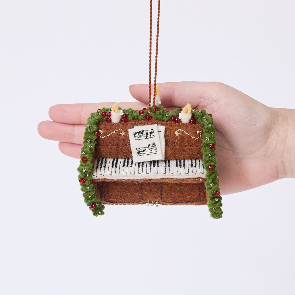 Craftspring’s Christmas Piano Ornament displayed in front of a hand to show its size — handmade from sustainable wool with a candle, evergreen garland, and embroidered sheet music, ethically crafted by artisans with intricate beaded and embroidered details.