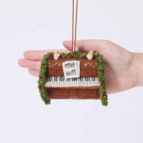 Craftspring’s Christmas Piano Ornament displayed in front of a hand to show its size — handmade from sustainable wool with a candle, evergreen garland, and embroidered sheet music, ethically crafted by artisans with intricate beaded and embroidered details.
