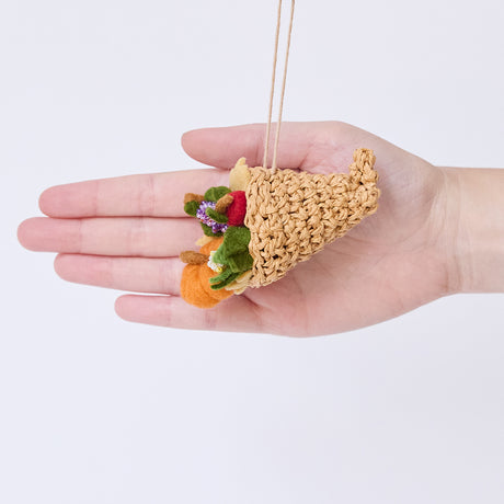 Craftspring’s Cornucopia ornament held in front of a hand to show its size—handmade from sustainable wool with a crocheted raffia cornucopia filled with felt and beaded vegetables and fruit, ethically crafted by artisans using sustainable wool.