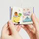 Hand-felted ornament shaped like a miniature crafting book, featuring three embroidered and appliquéd pages with illustrations of a pottery wheel, origami crane, knitting needles, and weaving loom. Made from merino-blend wool, this ornament includes a hanging string and measures approximately 2.5 inches tall. A charming tribute to handmade artistry and creative expression.