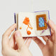 Hand-felted ornament shaped like a miniature crafting book, featuring three embroidered and appliquéd pages with illustrations of a pottery wheel, origami crane, knitting needles, and weaving loom. Made from merino-blend wool, this ornament includes a hanging string and measures approximately 2.5 inches tall. A charming tribute to handmade artistry and creative expression.