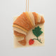 Hand-felted ornament of a golden-brown croissant resting on a pink felt napkin embroidered with a strawberry. Crafted from merino-blend wool with detailed stitching to mimic flaky layers, this ornament includes a hanging string and measures approximately 3.75 inches wide. A charming tribute to French pastry and cozy café mornings.