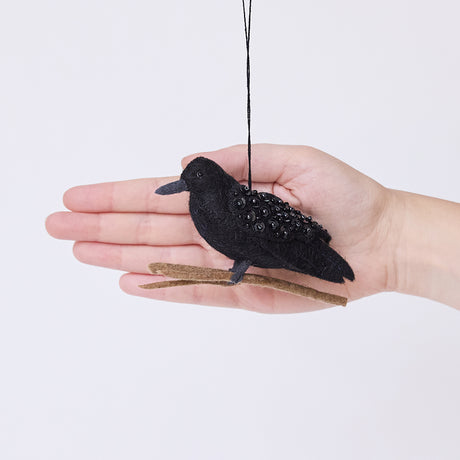 Craftspring’s Crow ornament held in front of a hand to show its size—handmade from sustainable wool with shimmering beaded wings, perched on a branch, perfect for Halloween, harvest decorating, and holiday gifting, ethically crafted by artisans using sustainable wool.