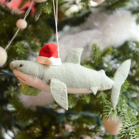 Craftspring’s Jolly Santa Shark with Gift Ornament — a cheerful shark wearing a Santa hat and holding a gift, with beaded and embroidered holiday details — displayed on a glowing Christmas tree, bringing humorous, joy-filled warmth to festive decorating and holiday gifting, ethically crafted by artisans using sustainable wool.