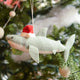 Craftspring’s Jolly Santa Shark with Gift Ornament — a cheerful shark wearing a Santa hat and holding a gift, with beaded and embroidered holiday details — displayed on a glowing Christmas tree, bringing humorous, joy-filled warmth to festive decorating and holiday gifting, ethically crafted by artisans using sustainable wool.