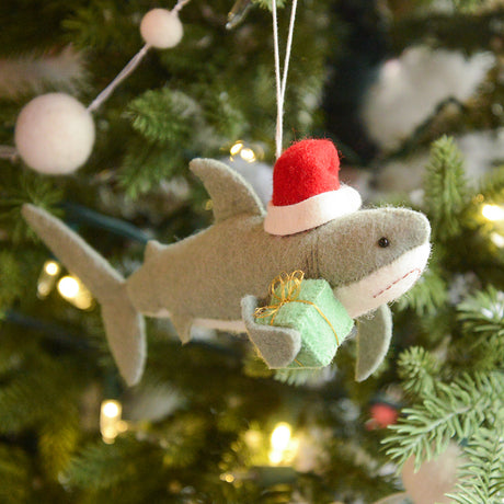 Craftspring’s Jolly Santa Shark with Gift Ornament — a cheerful shark wearing a Santa hat and holding a gift, with beaded and embroidered holiday details — displayed on a glowing Christmas tree, bringing humorous, joy-filled warmth to festive decorating and holiday gifting, ethically crafted by artisans using sustainable wool.