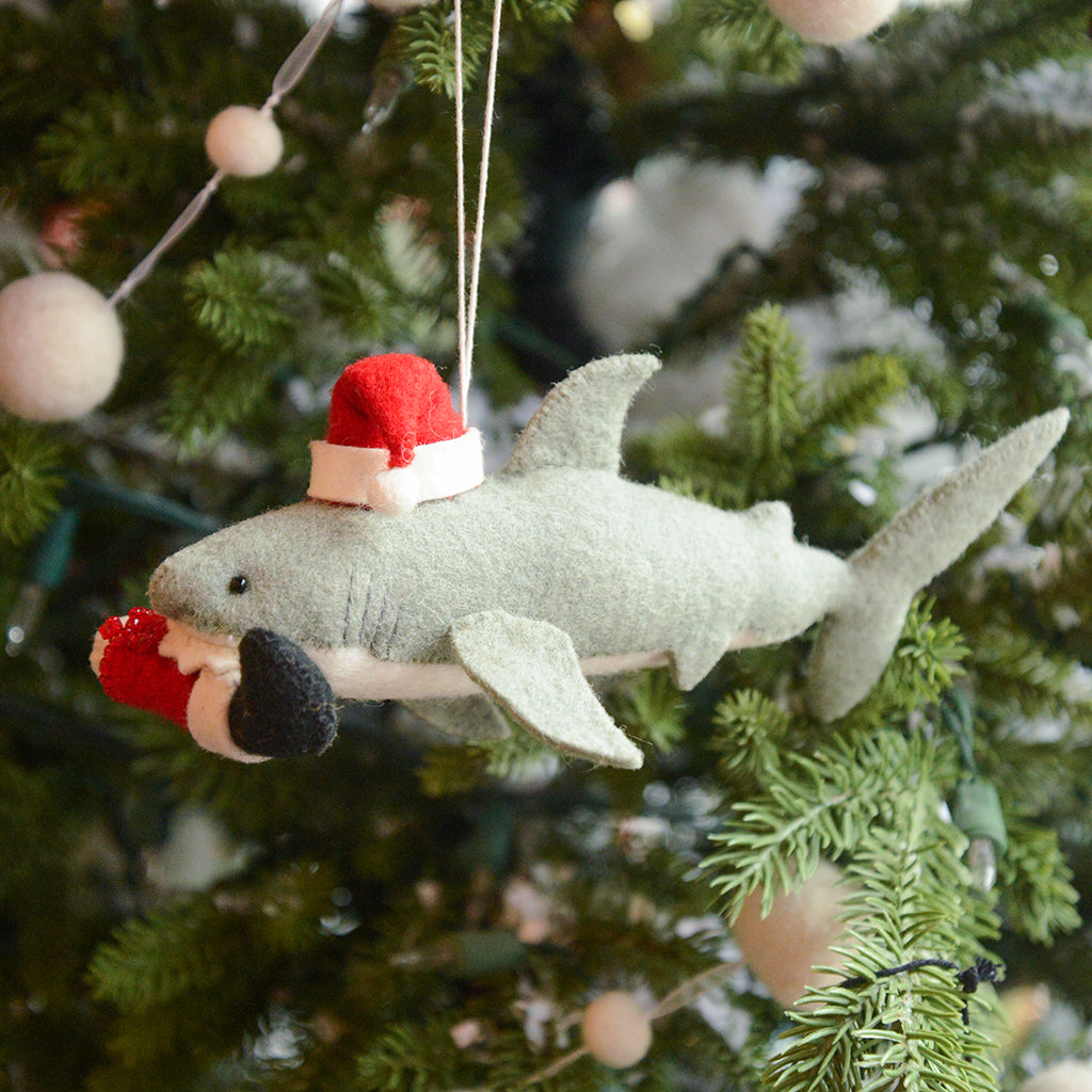 Craftspring’s Jolly Santa Shark with Santa Leg Ornament — a playful shark wearing a Santa hat and holding Santa’s leg in its mouth, with beaded and embroidered holiday details — displayed on a glowing Christmas tree, bringing humorous, joy-filled warmth to festive decorating and holiday gifting, ethically crafted by artisans using sustainable wool.