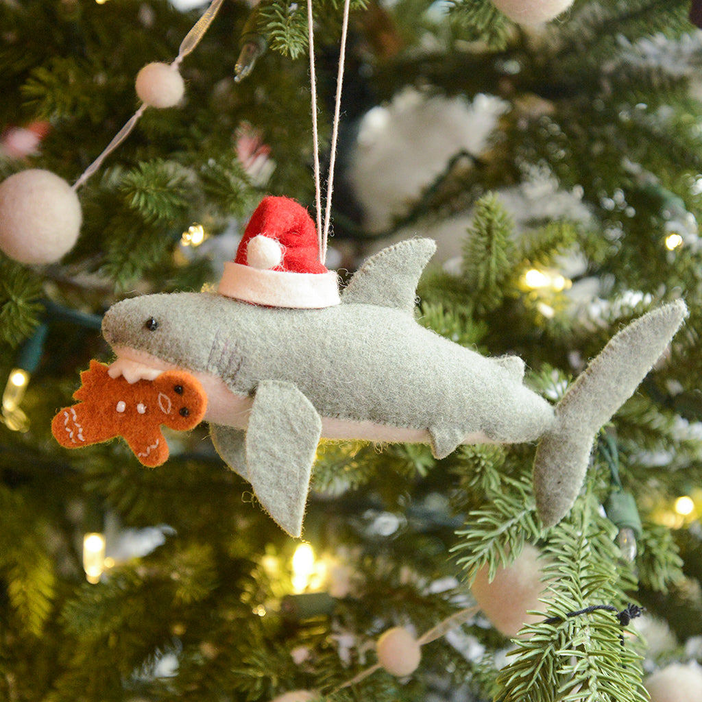 Craftspring’s Jolly Santa Shark with Gingerbread Ornament — a playful shark holding a gingerbread man in its teeth with beaded and embroidered holiday details — displayed on a glowing Christmas tree, bringing humorous, joy-filled warmth to festive decorating and holiday gifting, ethically crafted by artisans using sustainable wool.