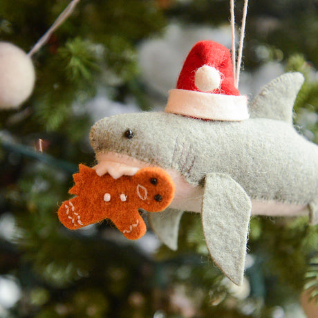 Craftspring’s Jolly Santa Shark with Gingerbread Ornament — a playful shark holding a gingerbread man in its teeth with beaded and embroidered holiday details — displayed on a glowing Christmas tree, bringing humorous, joy-filled warmth to festive decorating and holiday gifting, ethically crafted by artisans using sustainable wool.