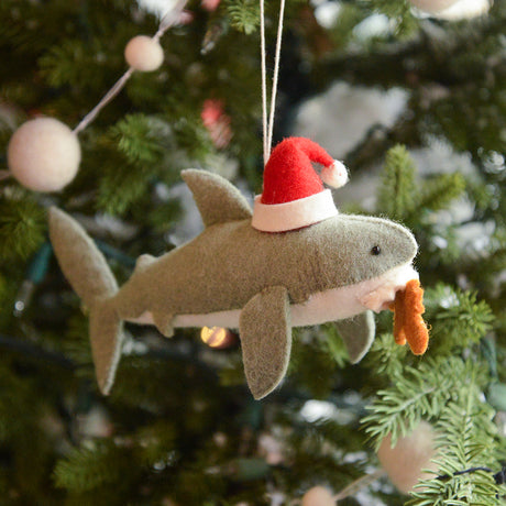 Craftspring’s Jolly Santa Shark with Gingerbread Ornament — a playful shark holding a gingerbread man in its teeth with beaded and embroidered holiday details — displayed on a glowing Christmas tree, bringing humorous, joy-filled warmth to festive decorating and holiday gifting, ethically crafted by artisans using sustainable wool.