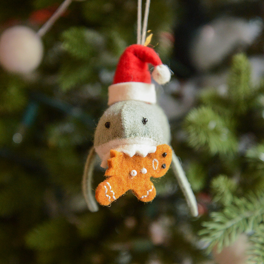 Craftspring’s Jolly Santa Shark with Gingerbread Ornament — a playful shark holding a gingerbread man in its teeth with beaded and embroidered holiday details — displayed on a glowing Christmas tree, bringing humorous, joy-filled warmth to festive decorating and holiday gifting, ethically crafted by artisans using sustainable wool.