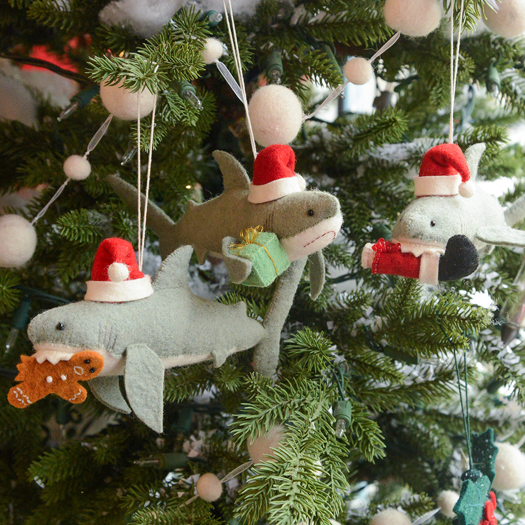 Craftspring’s Jolly Santa Shark with Gingerbread Ornament — featuring beaded and embroidered details and a gingerbread man clasped in its toothy grin — shown alongside two other holiday shark ornaments on a twinkling Christmas tree, creating a whimsical, gift-ready scene full of Christmas cheer and heirloom-quality charm, ethically crafted by artisans using sustainable wool.