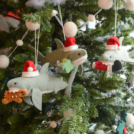 Craftspring’s Jolly Santa Shark with Gingerbread Ornament — featuring beaded and embroidered details and a gingerbread man clasped in its toothy grin — shown alongside two other holiday shark ornaments on a twinkling Christmas tree, creating a whimsical, gift-ready scene full of Christmas cheer and heirloom-quality charm, ethically crafted by artisans using sustainable wool.