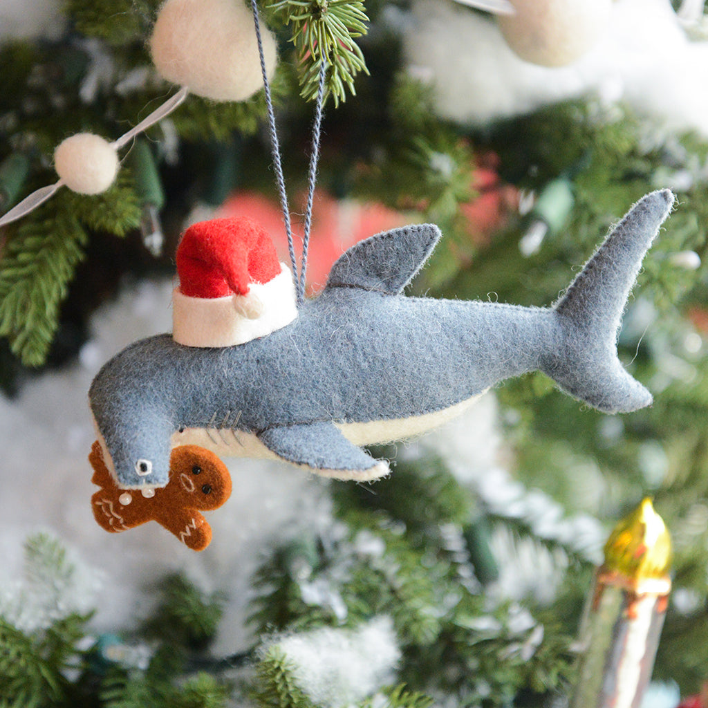 Craftspring’s Take a Bite Out of Christmas Hammerhead Ornament — a festive hammerhead shark wearing a Santa hat and munching a gingerbread man, detailed with beaded and embroidered holiday accents — displayed on a glowing Christmas tree, adding humorous charm, heirloom-quality warmth, and gift-ready Christmas cheer, ethically crafted by artisans using sustainable wool.