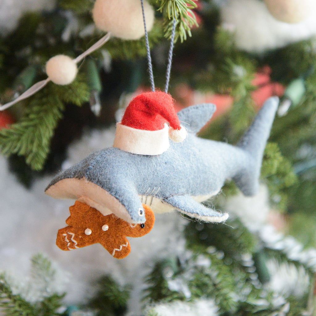 Craftspring’s Take a Bite Out of Christmas Hammerhead Ornament — a festive hammerhead shark wearing a Santa hat and munching a gingerbread man, detailed with beaded and embroidered holiday accents — displayed on a glowing Christmas tree, adding humorous charm, heirloom-quality warmth, and gift-ready Christmas cheer, ethically crafted by artisans using sustainable wool.
