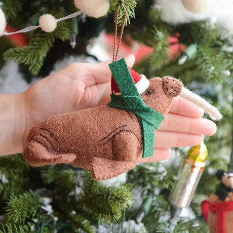 Craftspring’s Santa Walrus Ornament displayed in front of a hand to show its size — featuring a Santa hat, a holiday scarf, and soft embroidered details — an adorable, heirloom-quality piece perfect for joyful Christmas gifting, ethically crafted by artisans using sustainable wool.