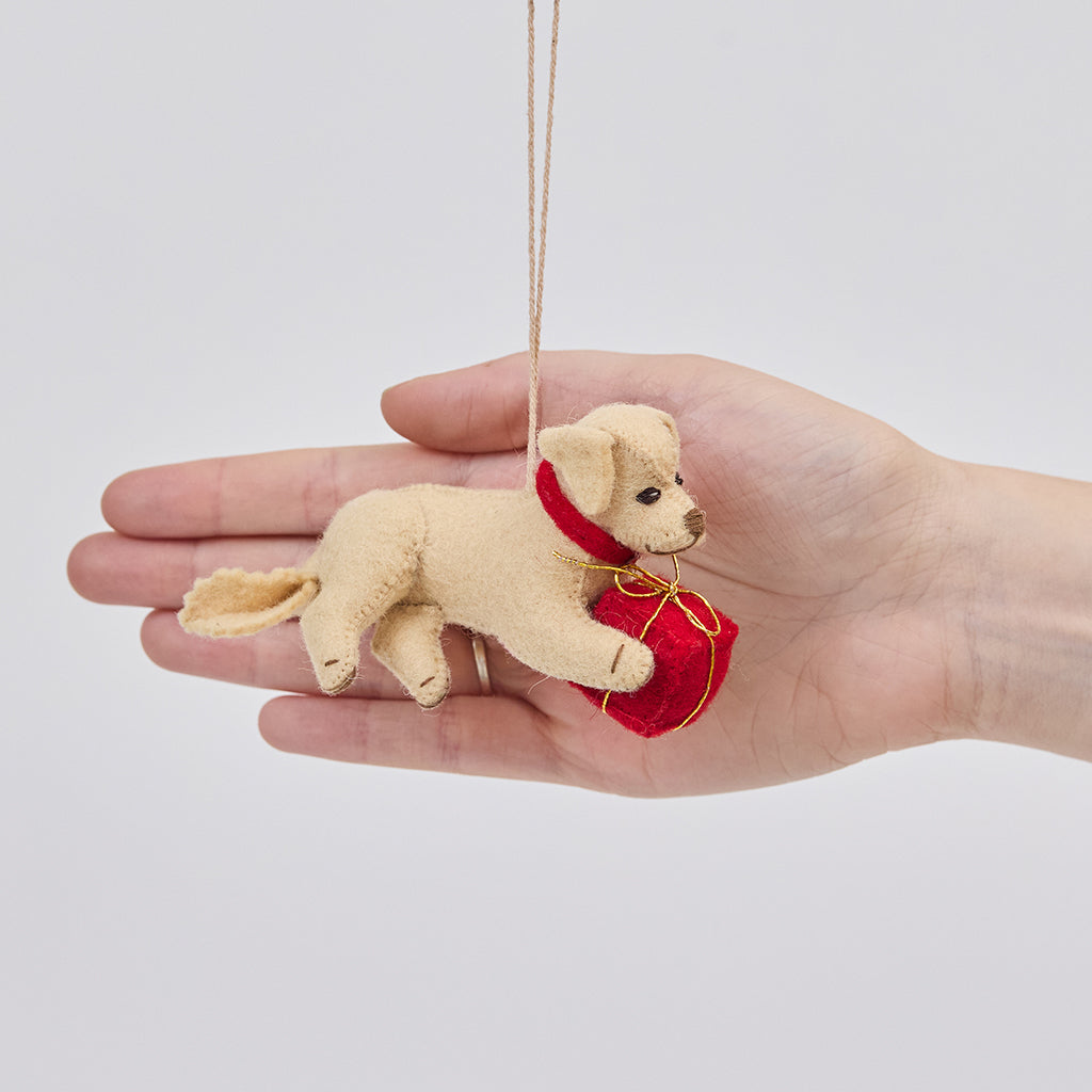 Handmade felt Dog with Gift ornament from Craftspring held in front of a hand to show its size—featuring a red present between its paws, with heirloom-quality stitching and festive charm, ethically crafted by artisans using sustainable wool.