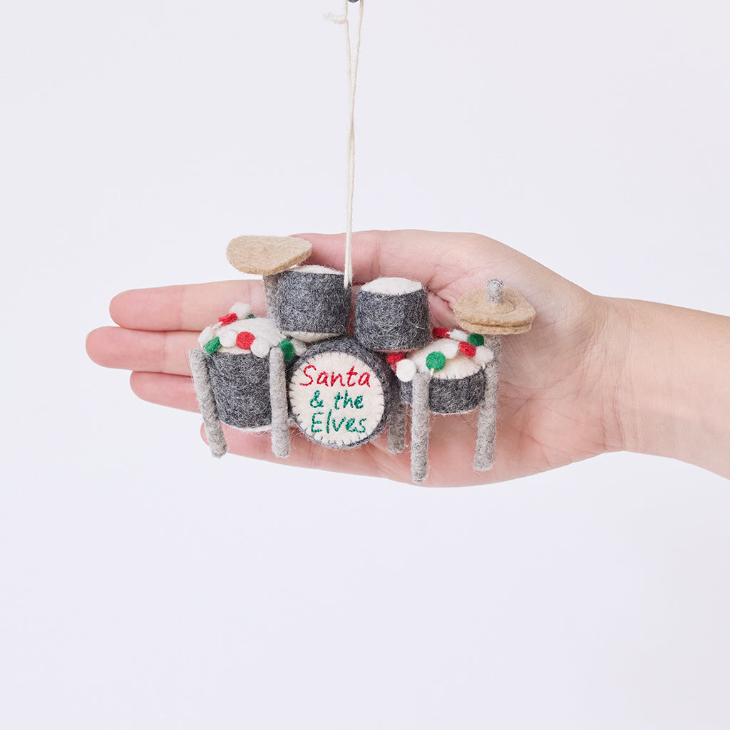 Craftspring’s Christmas Drum Set Ornament displayed in front of a hand to show its size — handmade from sustainable wool with embroidered “Santa and the Elves” band name and festive garland details, ethically crafted by artisans for heirloom holiday gifting.