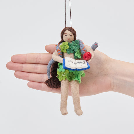 Craftspring’s Eve ornament held in front of a hand to show its size—handmade from sustainable wool with Eve holding a book of knowledge and an apple, wrapped in leaves with a serpent around her shoulders, ethically crafted by artisans using sustainable wool.