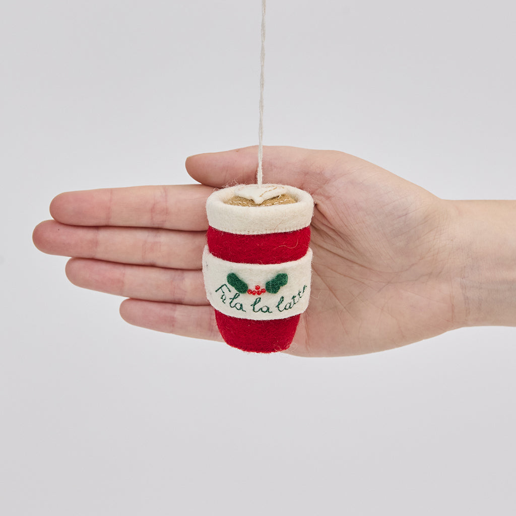Handmade felt Fa La La Latte ornament from Craftspring held in front of a hand to show its size—featuring detailed embroidery including “Fa la la latte” with holly, a Christmas tree, and a latte heart on top, ethically crafted by artisans using sustainable wool.