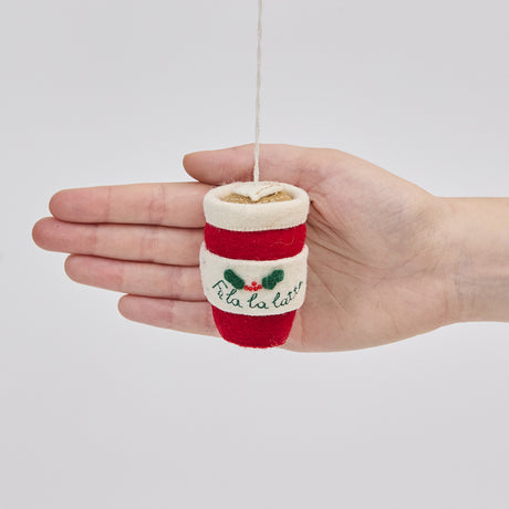 Handmade felt Fa La La Latte ornament from Craftspring held in front of a hand to show its size—featuring detailed embroidery including “Fa la la latte” with holly, a Christmas tree, and a latte heart on top, ethically crafted by artisans using sustainable wool.