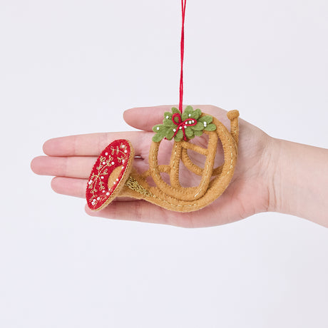 Craftspring’s French Horn Ornament displayed in front of a hand to show its size — handmade from sustainable wool with golden embroidery, beaded details, and mistletoe accents, ethically crafted by artisans.