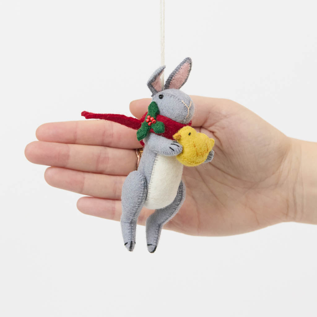Our Gray Holiday Bunny with Chick Ornament captures a heartwarming moment of gentle friendship. Wrapped in a cozy red scarf adorned with festive holly leaves and tiny red berries, this sweet gray bunny lovingly cradles a fluffy yellow chick, creating a scene that's full of warmth, comfort, and Christmas cheer.