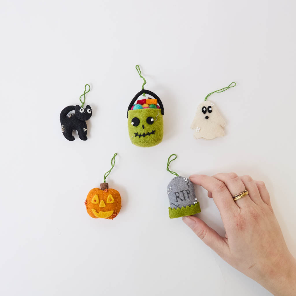 A handmade felt Halloween Mini Ornaments Set featuring five tiny spooky-season favorites: a black cat, candy-filled monster bucket, ghost, jack-o’-lantern, and RIP tombstone.