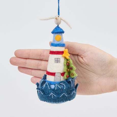 Handmade felt Holiday Lighthouse Christmas ornament from Craftspring held in front of a hand to show its size—featuring a seagull on top, crashing waves, and a Christmas tree, ethically crafted by artisans using sustainable wool.