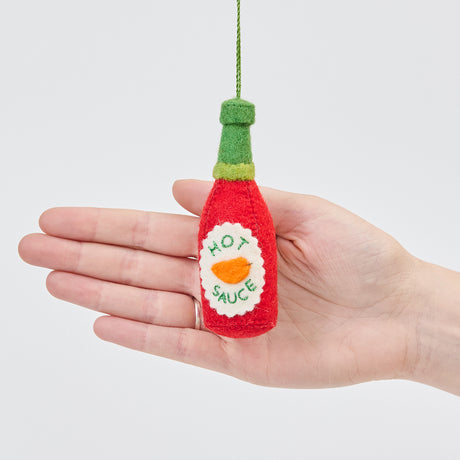Handmade felt Hot Sauce ornament from Craftspring, held in front of a hand to show its size—fun stocking stuffer for hot sauce fans, sustainably handmade by artisans using wool.