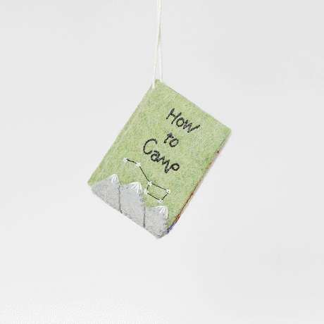 Handmade felt How to Camp Book ornament from Craftspring shown flipping through interior pages—featuring intricate beaded and embroidered details inspired by camping essentials, ethically crafted by artisans using sustainable wool.