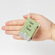 Hand-sewn felt ornament in the shape of a miniature book titled How to Camp, featuring embroidered pages with images of a tent, campfire, and fishing pole. Made from natural merino-blend wool, this whimsical ornament includes a hanging string and measures approximately 2.5 inches tall and 3 inches wide. With a hand for scale.